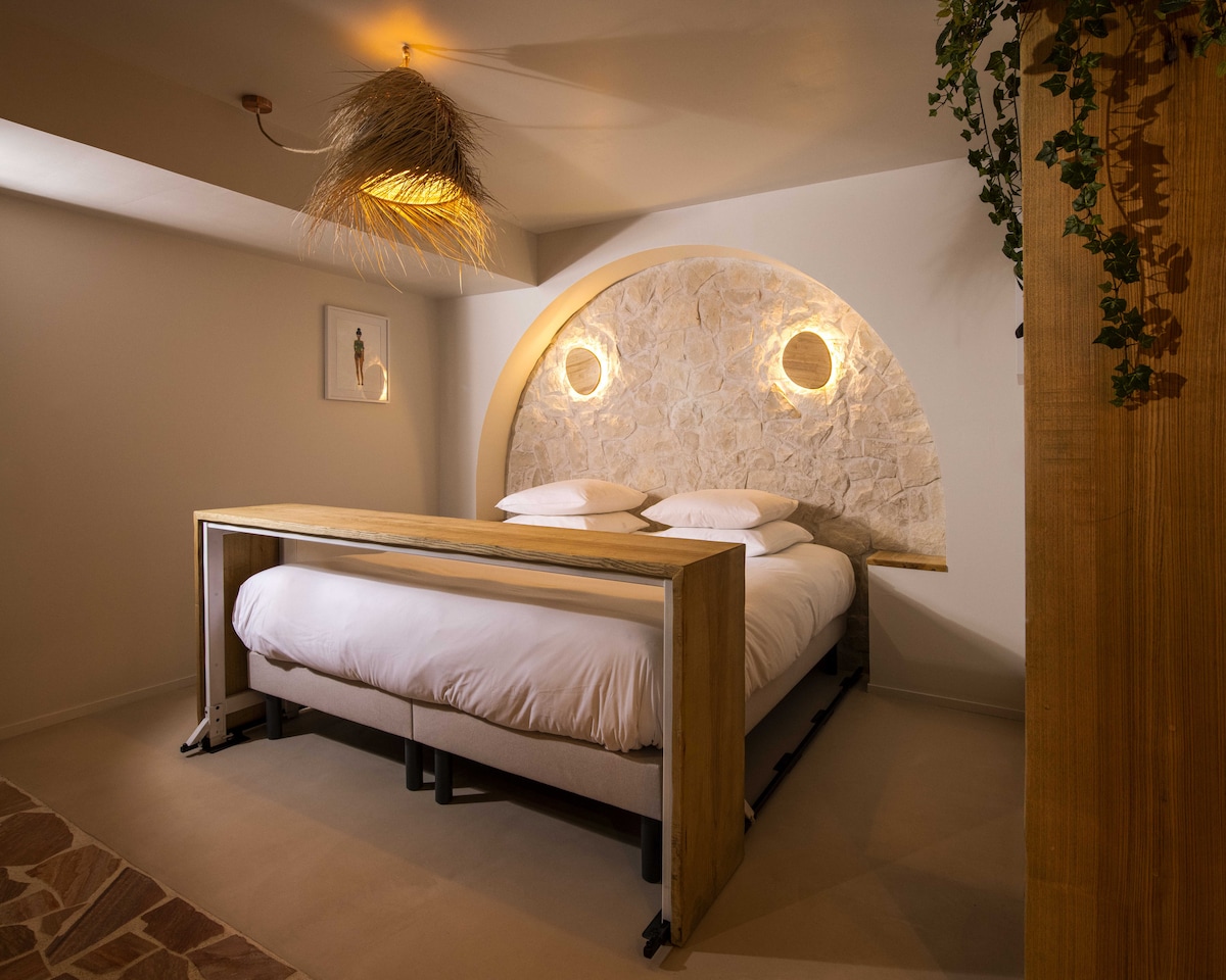 A spacious sleeping area features a large bed with white bedding, positioned against a decorative stone wall with circular light fixtures. Natural materials are incorporated through a wooden headboard and ceiling decor, while soft lighting contributes to a calming ambiance.