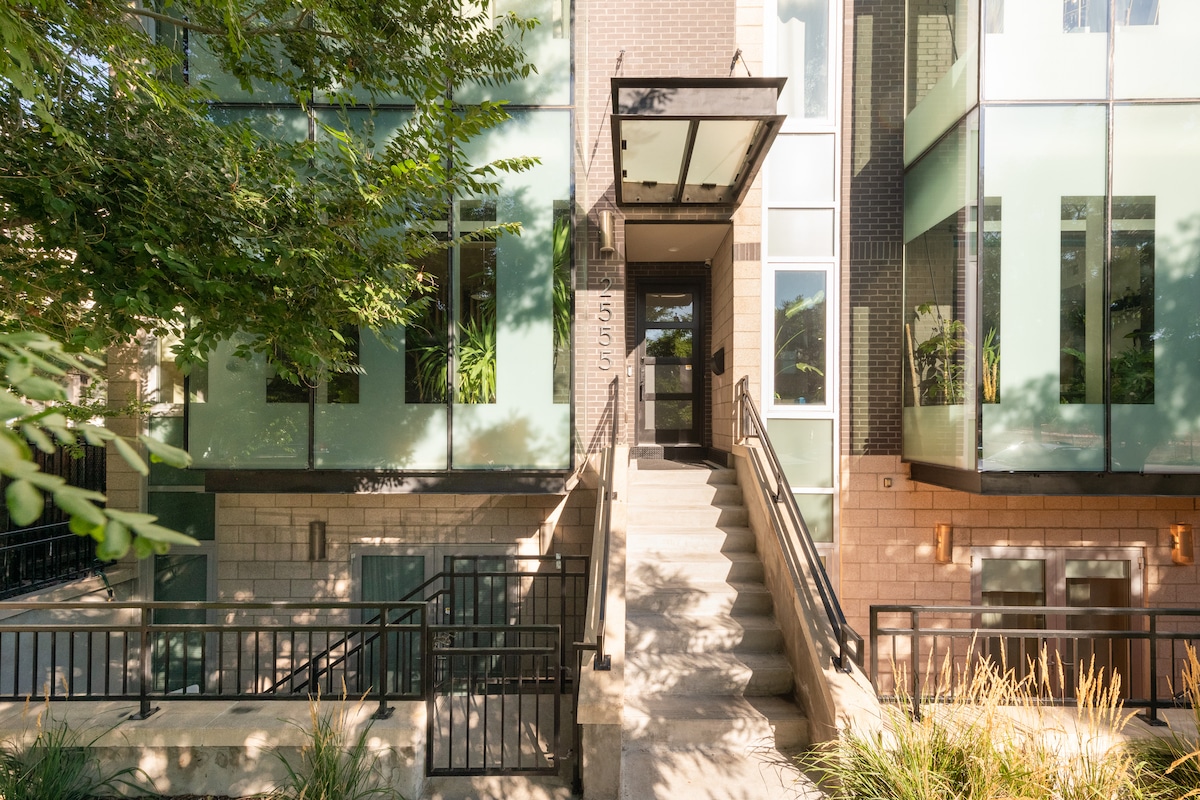 The entrance to a modern townhouse is visible, featuring a series of steps leading up to a glass door. Lush greenery surrounds the exterior, with large windows framed by a combination of brick and glass materials, emphasizing the contemporary design.