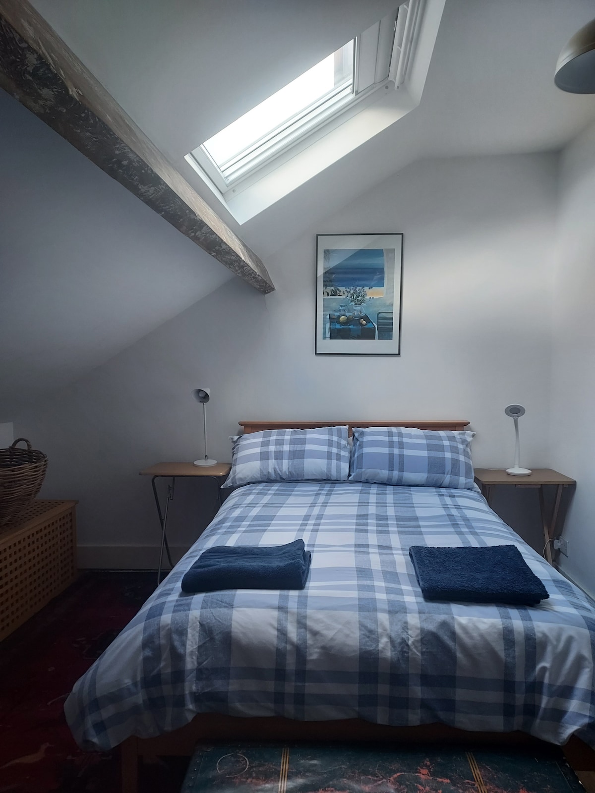 A cozy double room features a sloped ceiling with exposed beams. Natural light enters through a skylight, illuminating the plaid bedding. Two bedside tables are positioned on either side of the bed, each with a reading lamp. A woven basket adds a touch of warmth.