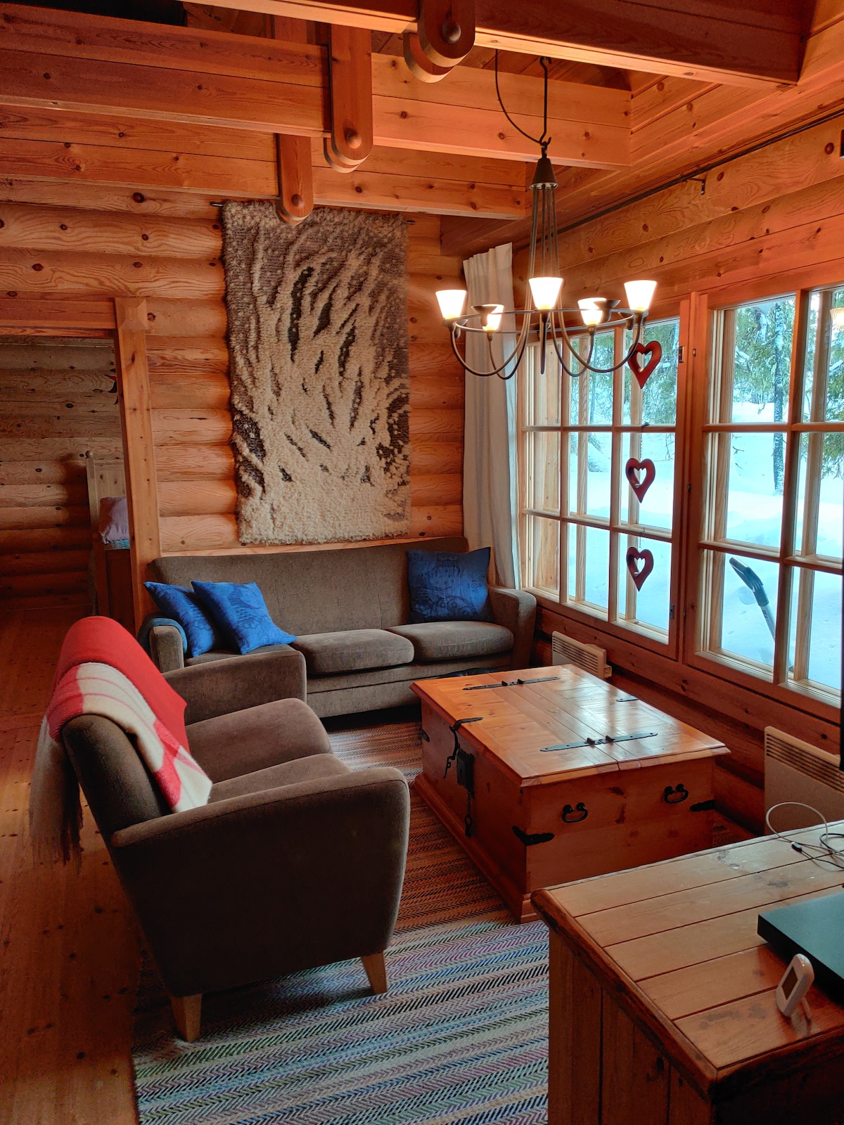 A living area featuring wooden walls and a colorful woven rug. Two sofas are positioned around a wooden coffee table. A chandelier with five lights hangs above, and large windows allow natural light to fill the space, revealing a snowy outdoor scene.