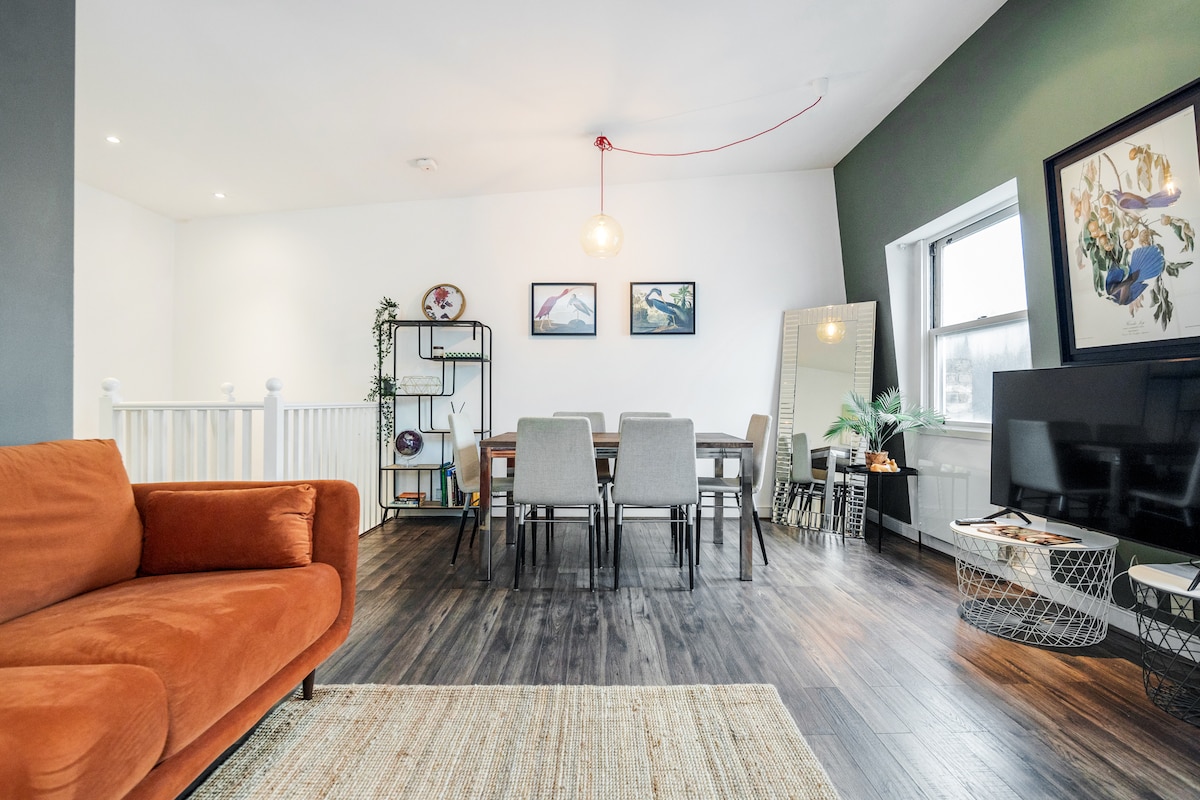 A spacious lounge area is visible, featuring a rust-colored couch and a dining table surrounded by six chairs. Natural light enters through a large window, illuminating the room. A large mirror and various decorative elements add visual interest to the inviting space.