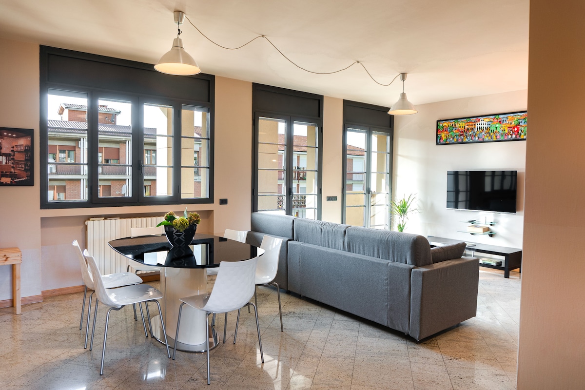 A modern living area features large windows allowing natural light to fill the space. A circular table with six white chairs is positioned near the windows, while a gray sofa faces a wall-mounted television. Decorative artwork is displayed above the sofa.