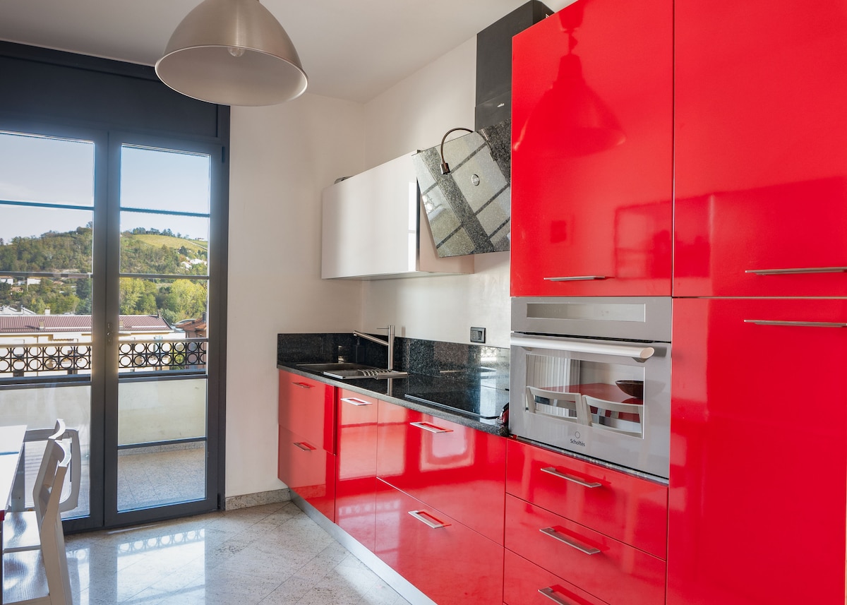 A modern kitchen features sleek red cabinetry complemented by black countertops. Stainless steel appliances are incorporated, and a large window offers views of the surrounding landscape. A dining table is visible in the adjacent area, enhancing the inviting atmosphere of the space.