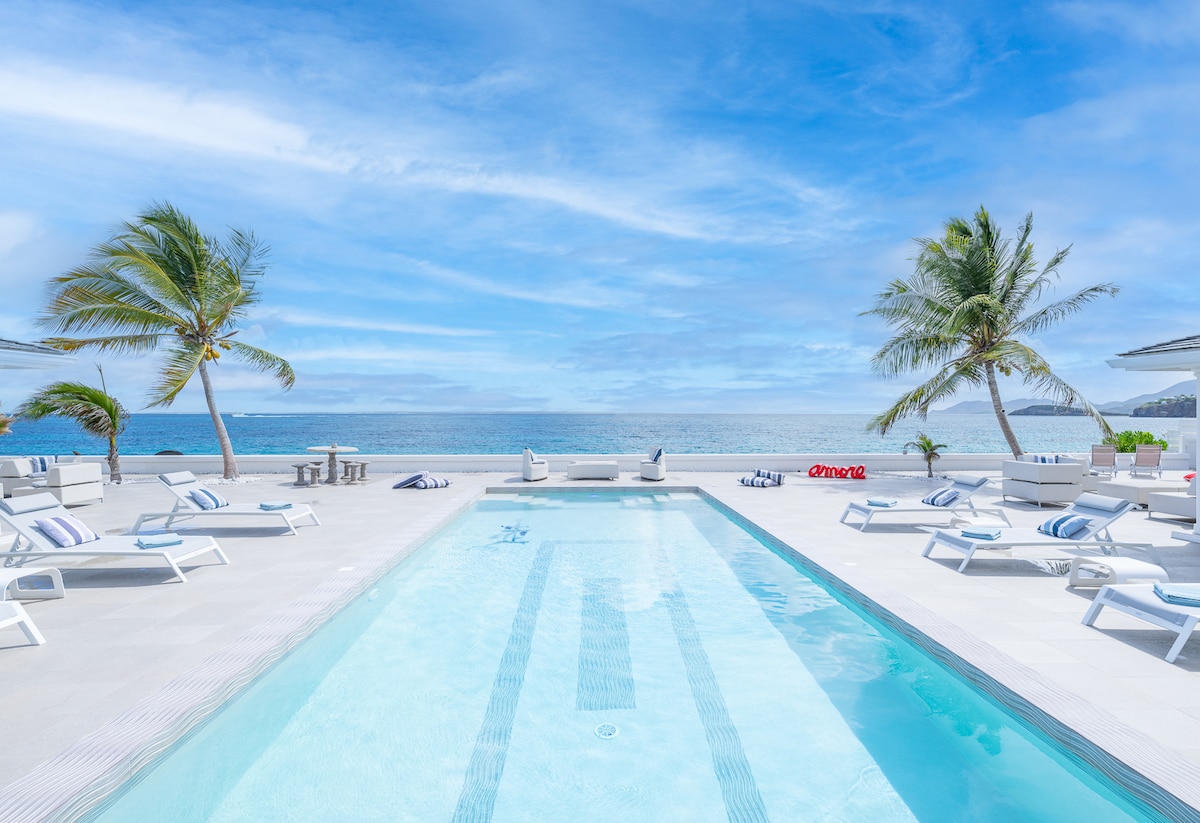A spacious pool area is bordered by white loungers, with palm trees gently swaying nearby. The clear blue water of the pool reflects the bright sky and ocean waves beyond, creating a serene outdoor setting.