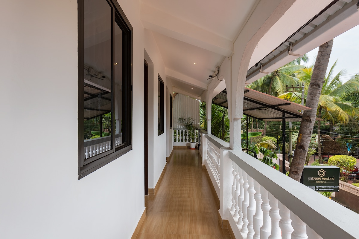 A covered corridor is seen, featuring smooth wooden flooring and white walls. Large windows allow natural light to filter in, providing views of the surrounding greenery, including palm trees. The corridor leads to additional areas of the property, enhancing accessibility.