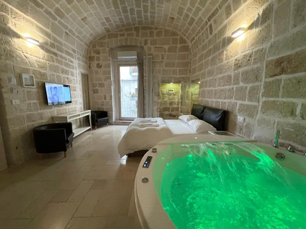 A spacious room features stone walls and a vaulted ceiling, creating a unique ambiance. A large jacuzzi with green lighting is positioned next to a comfortable bed. A flat-screen TV is mounted on the wall, and large windows provide natural light while offering a view of the outside.