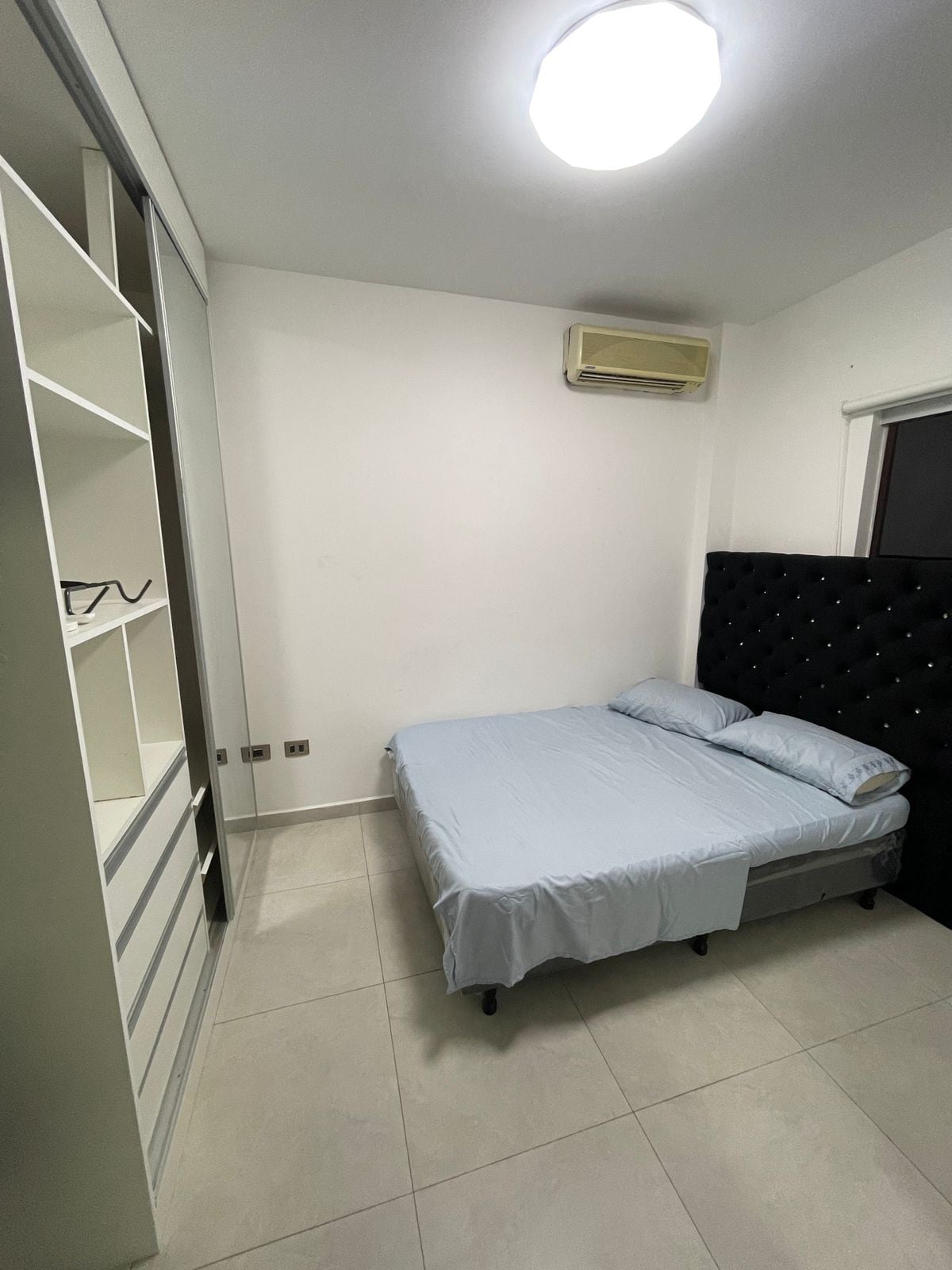 A compact bedroom features a bed with a tufted headboard and light blue bedding. A built-in wardrobe with open shelving and drawers is located on one wall, while a window with an air conditioning unit allows for airflow in the room.