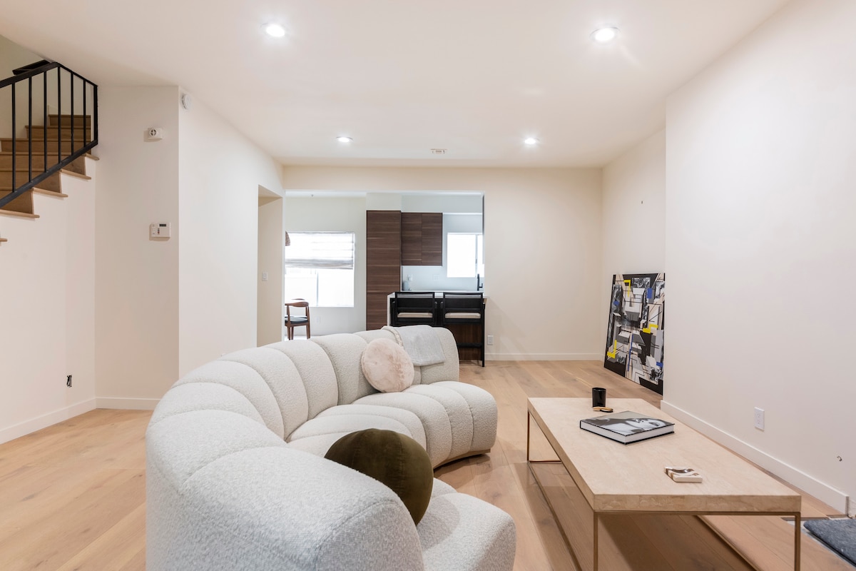 A spacious living area is depicted, featuring a curved sectional sofa in soft white fabric. A light wooden coffee table is centrally positioned, adorned with a candle and various magazines. Natural light enters through a nearby window, illuminating the minimalist decor.