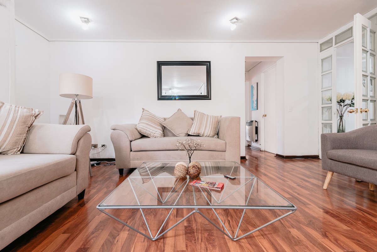 A living room features two light-colored sofas with patterned cushions, positioned around a modern glass coffee table. A decorative plant sits on the table, and a mirror reflects the space. Soft lighting creates a calm ambiance in the room.