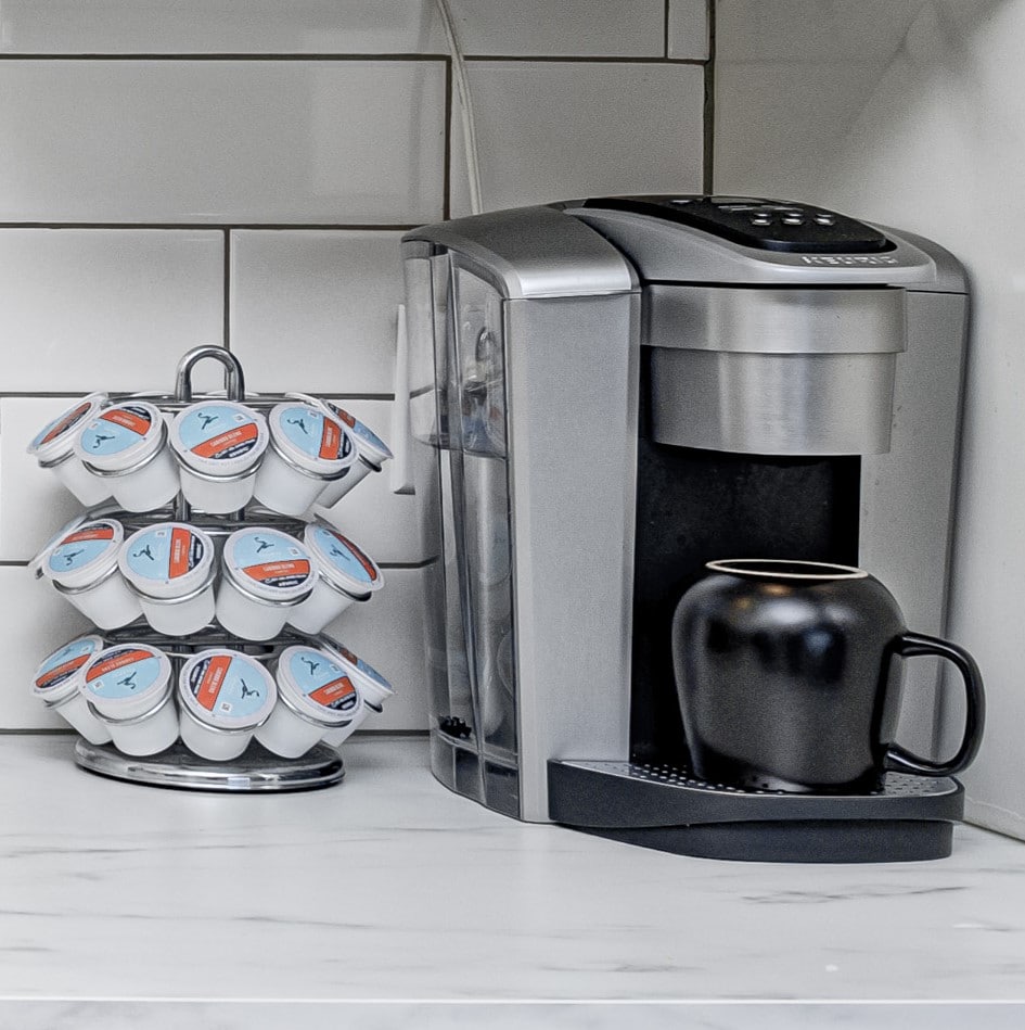 A sleek silver coffee maker is positioned beside a compact, circular holder filled with assorted coffee pods. A black ceramic mug rests near the coffee maker, ready to be filled. The countertop features light marble tones, creating a clean and modern look.