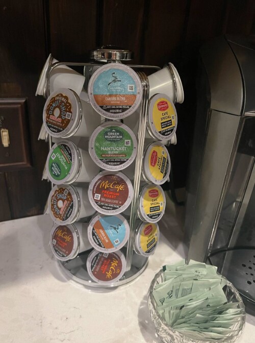 A coffee pod carousel is displayed on a countertop, showcasing a variety of flavored coffee pods organized in a circular holder. A small dish with sugar packets sits nearby, providing additional options for coffee preparation.