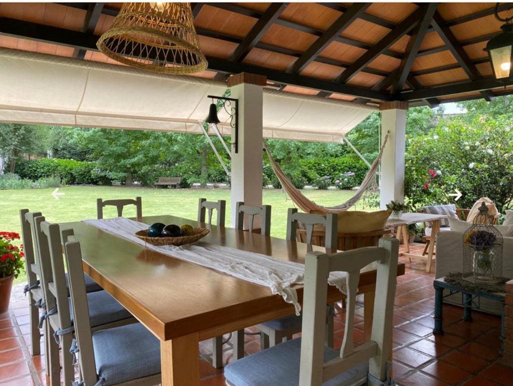 An outdoor dining area features a large wooden table surrounded by several light-colored chairs. A woven chandelier hangs overhead, and a hammock is positioned nearby. Lush greenery and flowering plants create a serene atmosphere, with natural light filtering through the open space.
