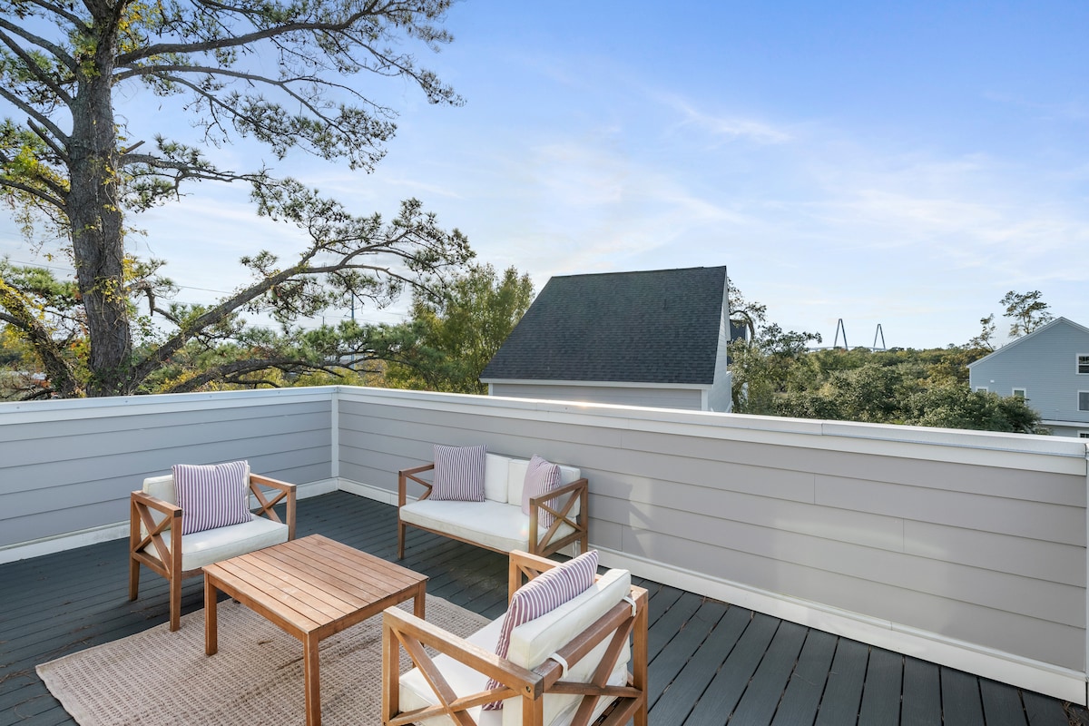 An outdoor rooftop patio is presented, featuring a wooden table surrounded by two armchairs and a loveseat with light cushions. A gentle breeze can be felt, complemented by views of neighboring rooftops and the lush trees in the surrounding area.