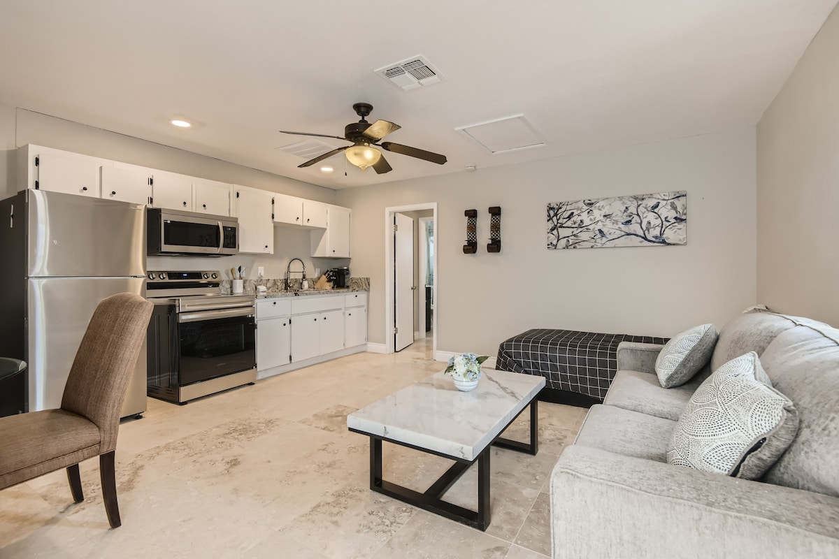 An open living area features a comfortable sofa and a stylish coffee table positioned in front of a wall-mounted television. A fully equipped kitchen with stainless steel appliances and white cabinets is visible in the background, complemented by a small dining area.