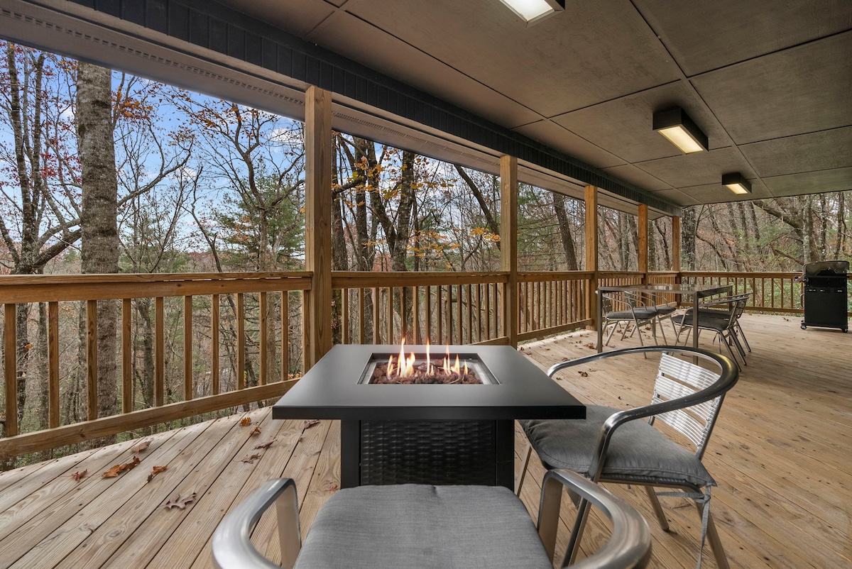 A spacious outdoor deck features a gas fire pit surrounded by seating. The deck offers views of the surrounding trees, with a gas grill positioned nearby. Natural light filters through the area, enhancing the inviting setting for relaxation.