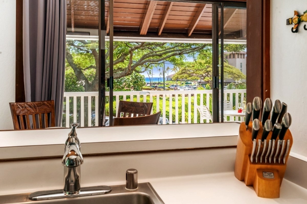 A view from the kitchen countertop showcases a bright outdoor space, featuring a sliding glass door that opens to a covered lanai with ocean views. A set of knives sits neatly in a wooden block beside the sink, with green trees visible beyond the lanai.
