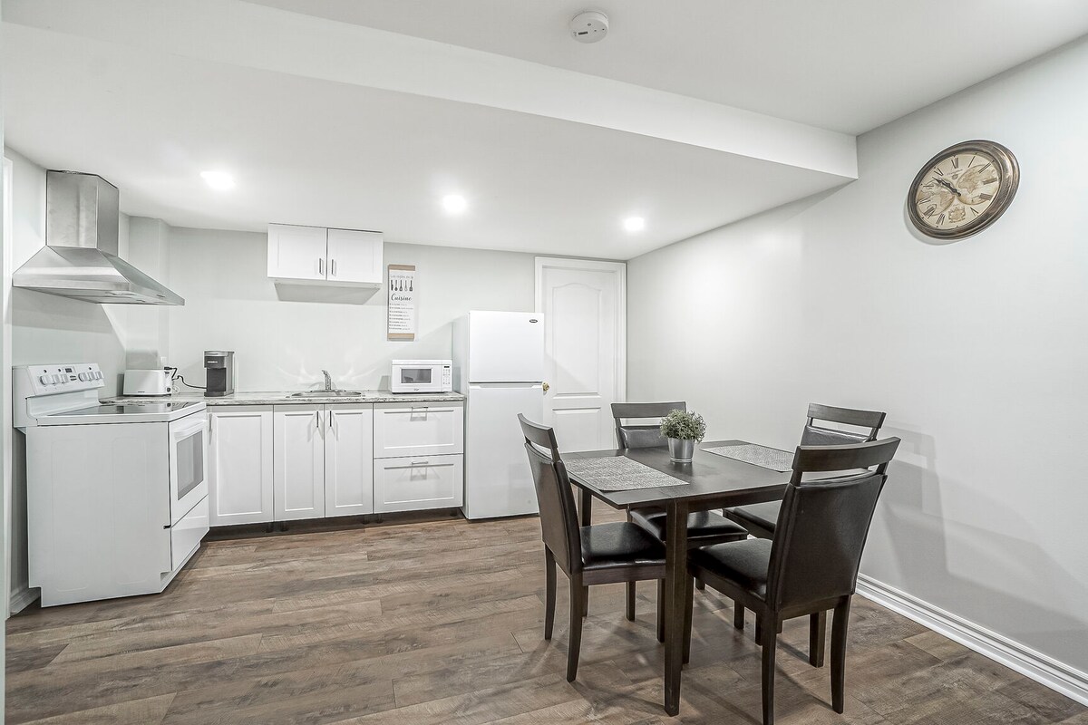 The kitchen and dining area are presented in an open layout, featuring modern appliances including a stove, refrigerator, and a coffee maker. A wooden dining table is set for four, complemented by dark chairs. Soft overhead lighting enhances the neutral-toned walls and flooring.