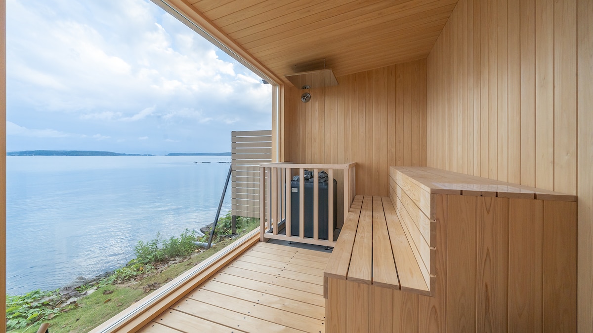 A wooden sauna interior is shown, featuring benches along the walls and a window that reveals a serene view of the water. Soft natural light filters in, highlighting the smooth wooden surfaces and creating a calming atmosphere.