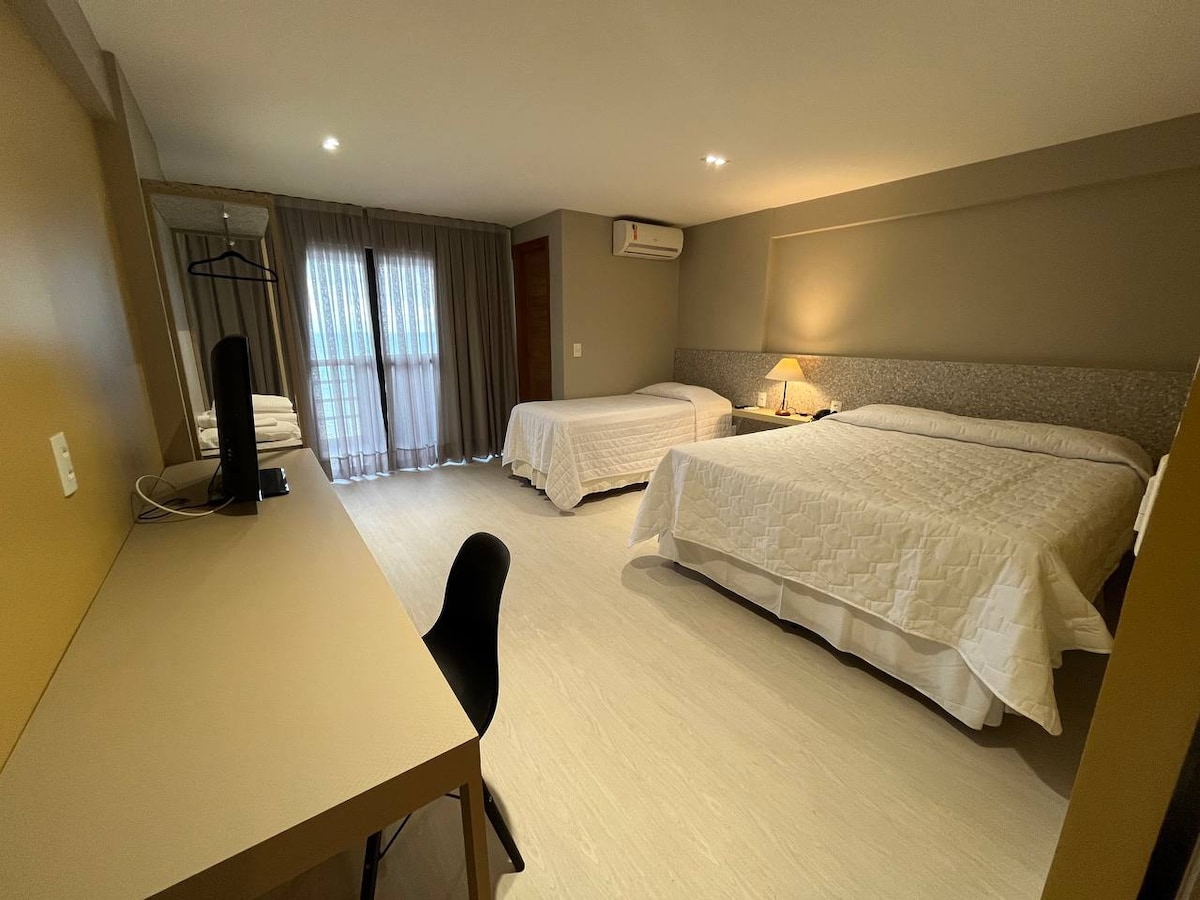 The spacious room features two large beds covered with white linens. A wooden desk sits beside a chair, while a window with light-filtering curtains allows natural light to enter. A closet with hangers is placed near the entry, enhancing the functional layout.