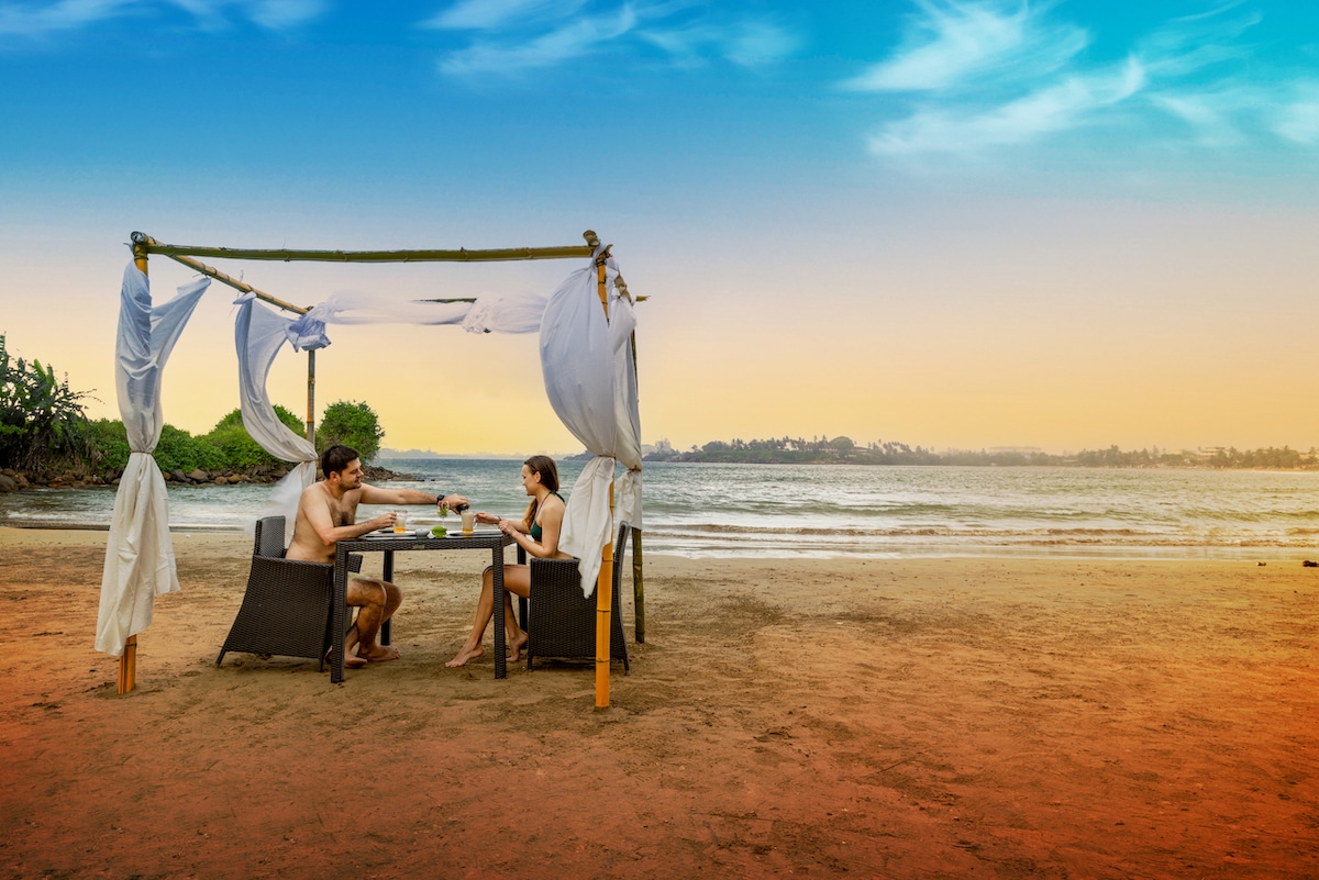 A cozy seating area features a bamboo structure adorned with flowing drapes along the beach, designed for intimate dining. Two chairs surround a small table, where refreshing beverages are present. The serene ocean and a colorful sunset create a tranquil backdrop.
