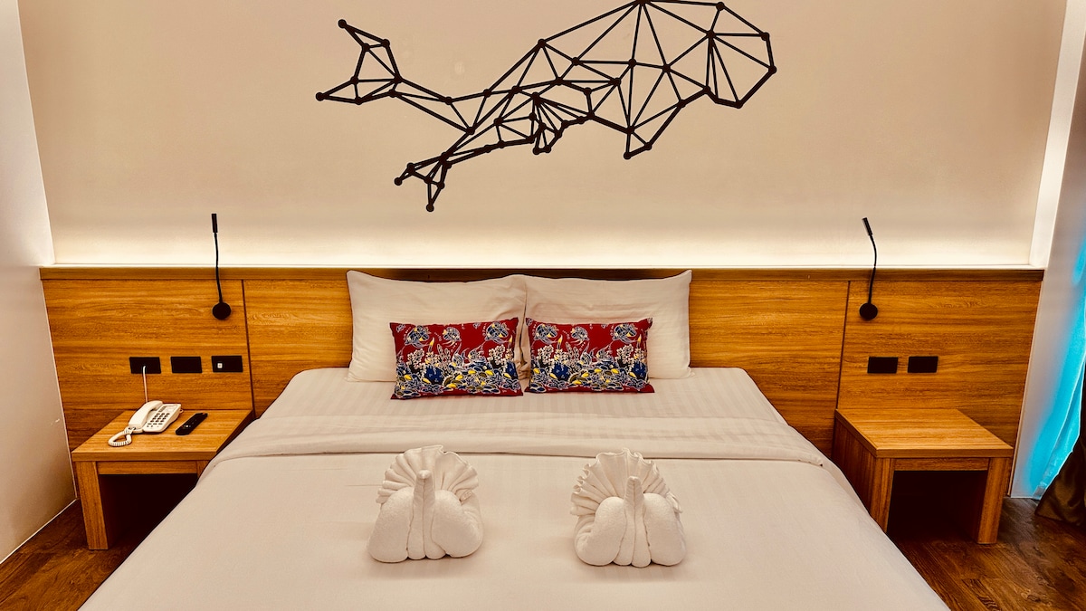 A spacious bed is centered beneath a geometric whale art piece. Two bedside tables are positioned on either side of the bed, complementing the wooden accents. Decorative towels are arranged in the shape of swans at the foot of the bed, creating a welcoming look.