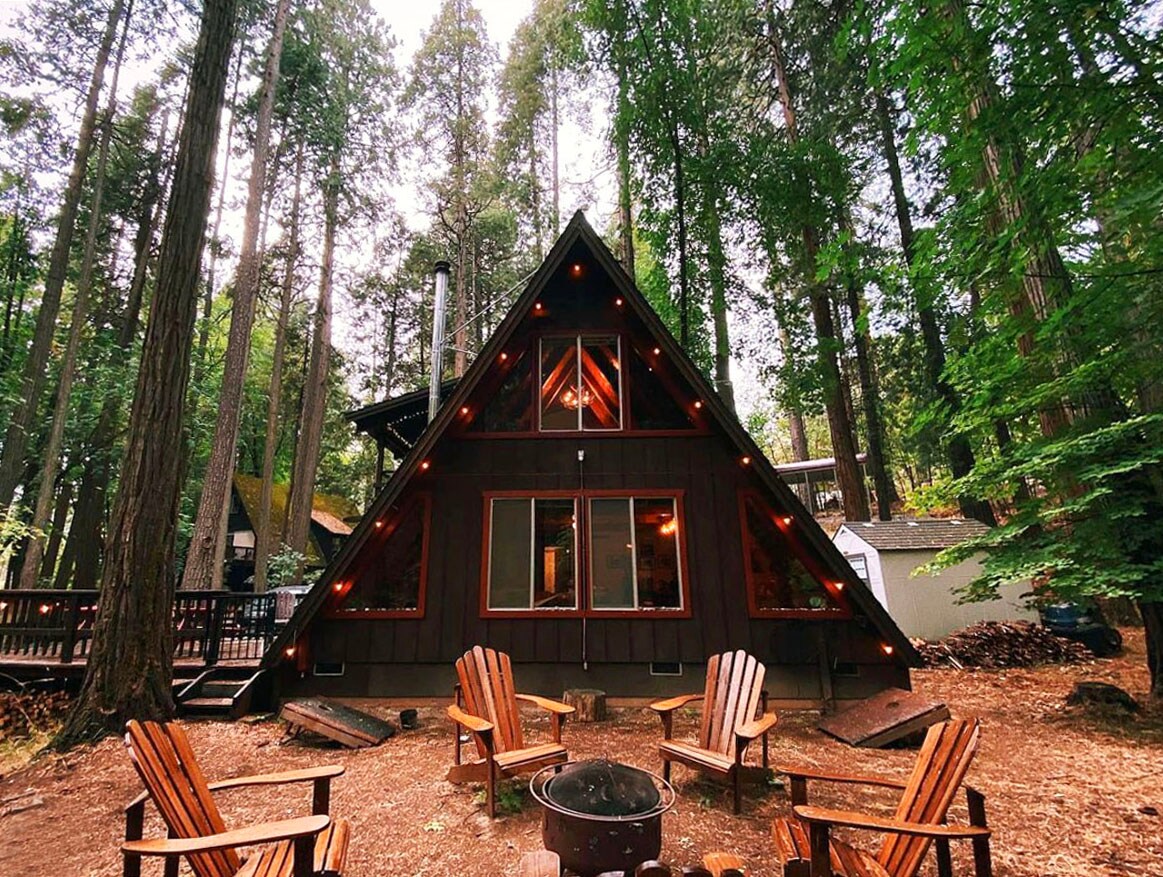 The A-frame cabin is surrounded by towering trees, featuring a welcoming façade with multiple large windows and an inviting entry. A circular arrangement of wooden chairs encircles a fire pit in the foreground, creating a cozy gathering space for relaxation.