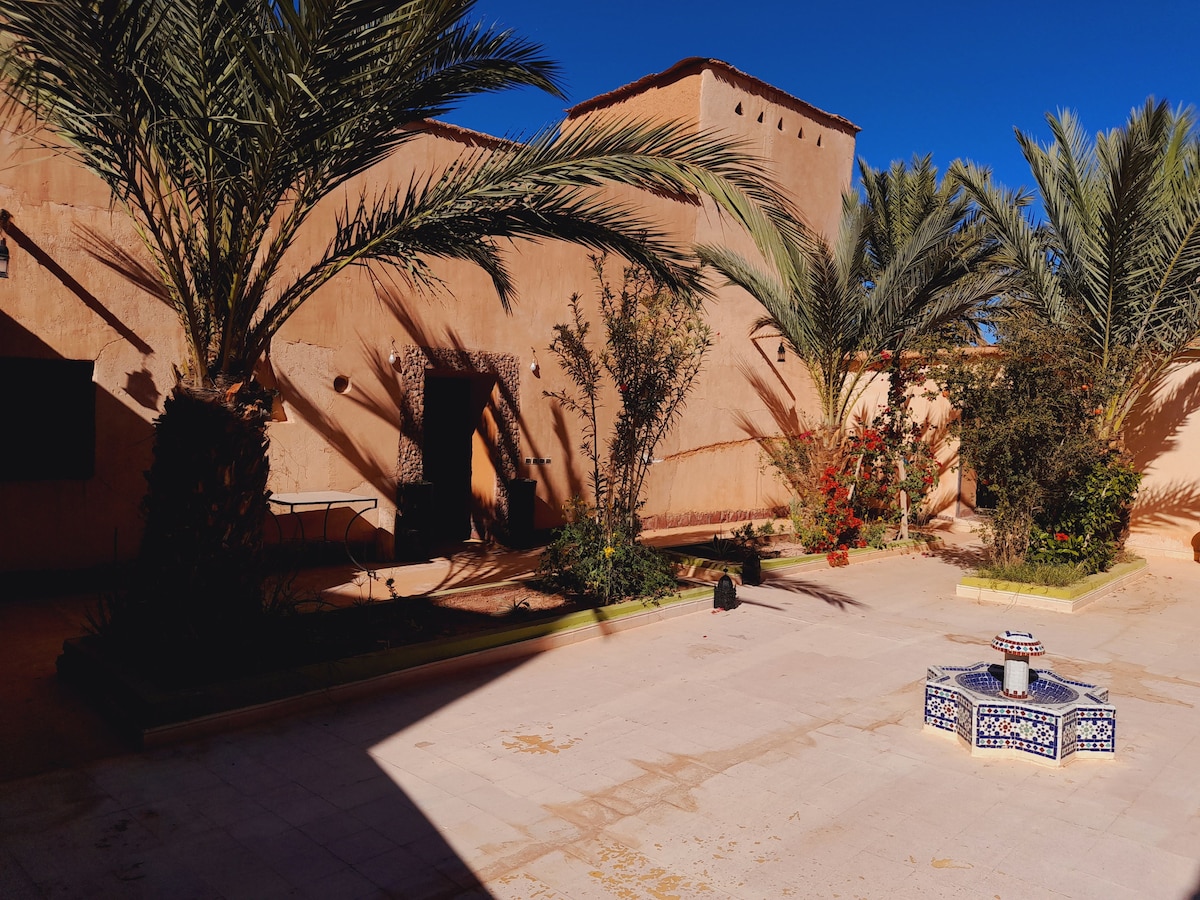A sunlit courtyard is surrounded by palm trees and lush greenery. A decorative fountain sits at the center, complemented by intricately patterned tiles. The earthy walls of the surrounding structures provide a warm backdrop, contributing to a peaceful atmosphere.