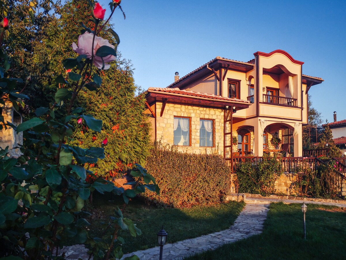 The two-story house is set among lush greenery, featuring a stone facade and a welcoming balcony. Sunlight bathes the exterior, highlighting the colorful garden filled with vibrant flowers. A path leads to the entrance, providing a glimpse of the inviting outdoor space.