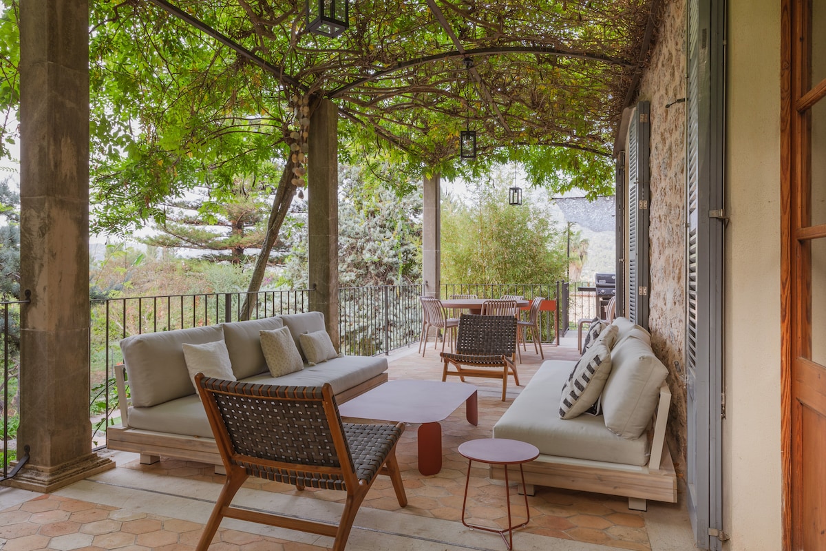 A shaded terrace is furnished with comfortable seating and a small accent table. Lush green vines create a natural canopy overhead, complementing the surrounding plants. A dining area is also present, providing a peaceful space for relaxation or meals while enjoying views of the garden.