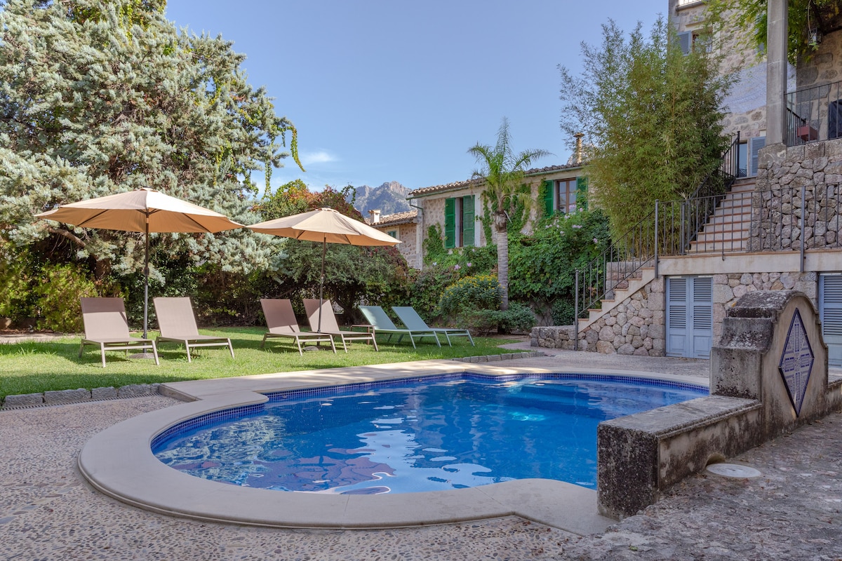 A serene outdoor space features a circular pool surrounded by lush greenery. Four sun loungers are positioned nearby, accompanied by two large umbrellas for shade. The stone steps lead up to the home, which is partially obscured by trees, creating a private atmosphere.