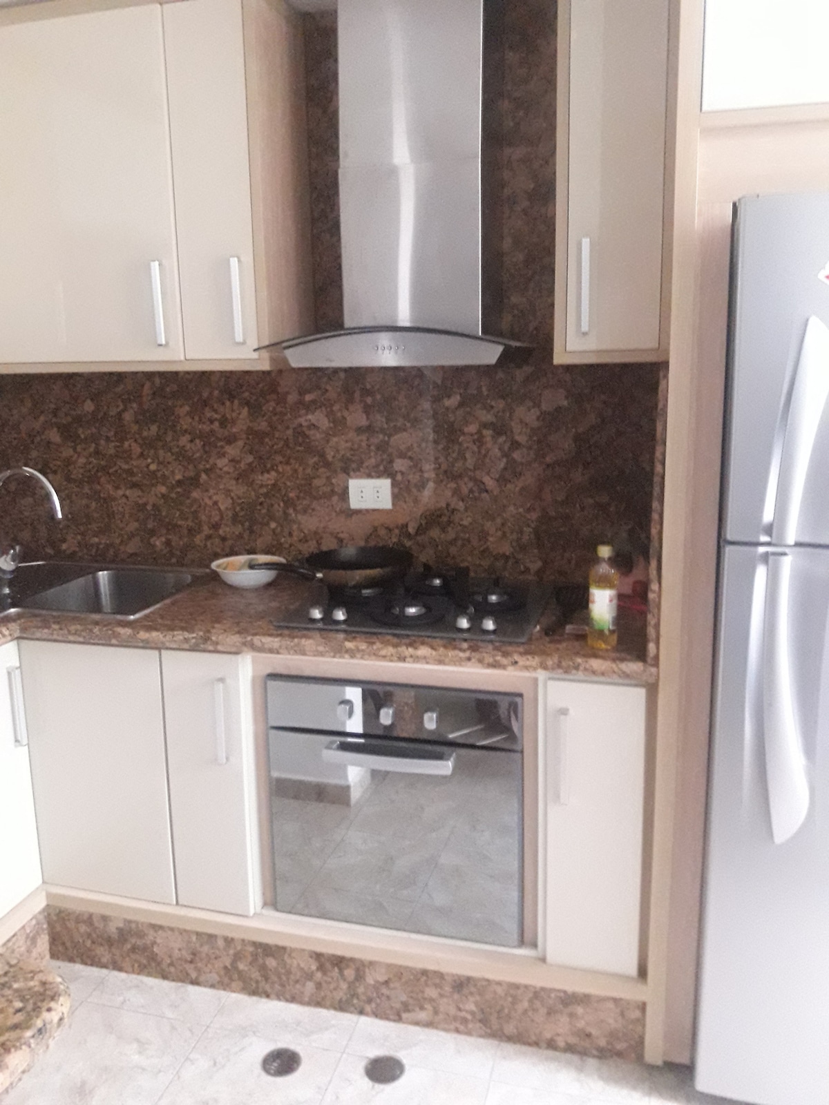 A compact kitchen area is presented, featuring sleek cabinetry with a light finish. A stainless steel refrigerator stands beside a built-in oven and five-burner cooktop. The countertop displays a patterned stone surface, and a sink is positioned under a window with ample natural light.