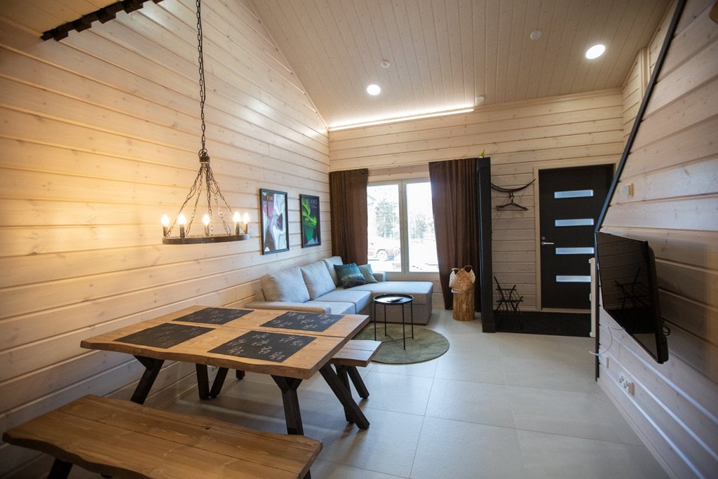 A living area is presented with wooden walls and a light wood ceiling. A spacious sofa sits alongside a round rug, adjacent to a dining table crafted from wood. A modern fixture with multiple bulbs hangs overhead, while a flat-screen TV is mounted on the wall.