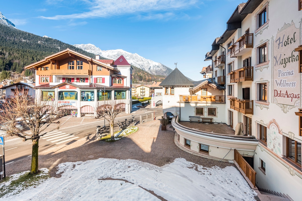 The image captures a scenic view of a hotel's exterior, flanked by picturesque mountains in the background. Snow covers parts of the landscape, while trees line the pathway. The building's architecture reflects a blend of traditional and modern styles, providing a welcoming atmosphere.