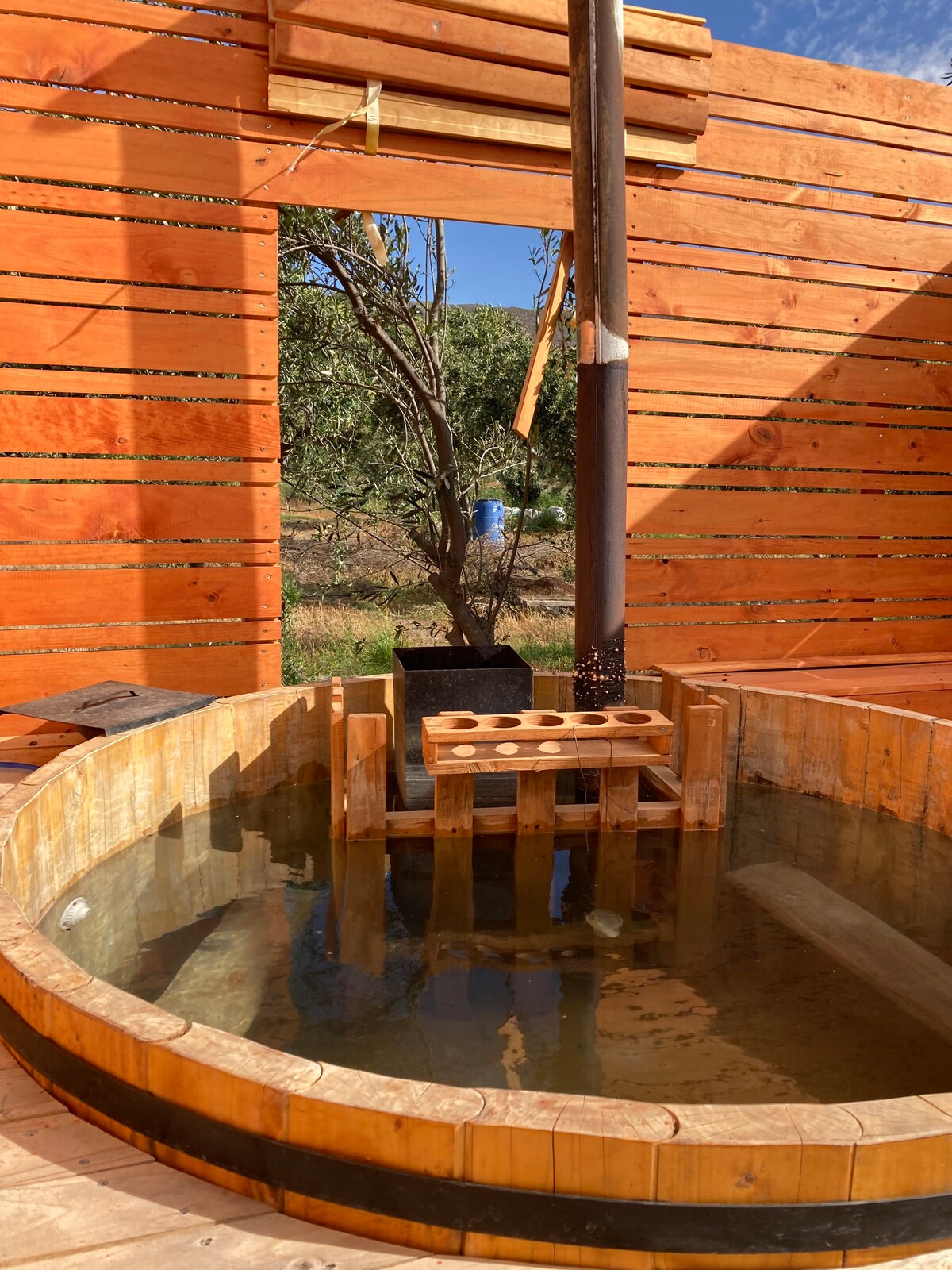 A wooden hot tub is featured within a rustic enclosure, surrounded by wooden slats that create a warm atmosphere. Clear water fills the round tub, while a simple wooden structure for seating is visible beside it. A glimpse of greenery is seen through the opening.