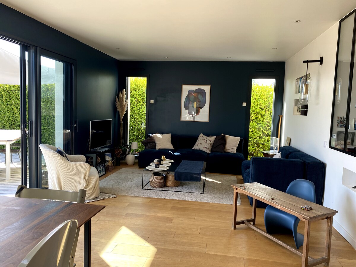 A modern living area features a dark wall design and large windows that bring in natural light. A comfortable sectional sofa is paired with a small coffee table, while a textured rug adds warmth to the space. A sleek television is positioned near a decorative plant.