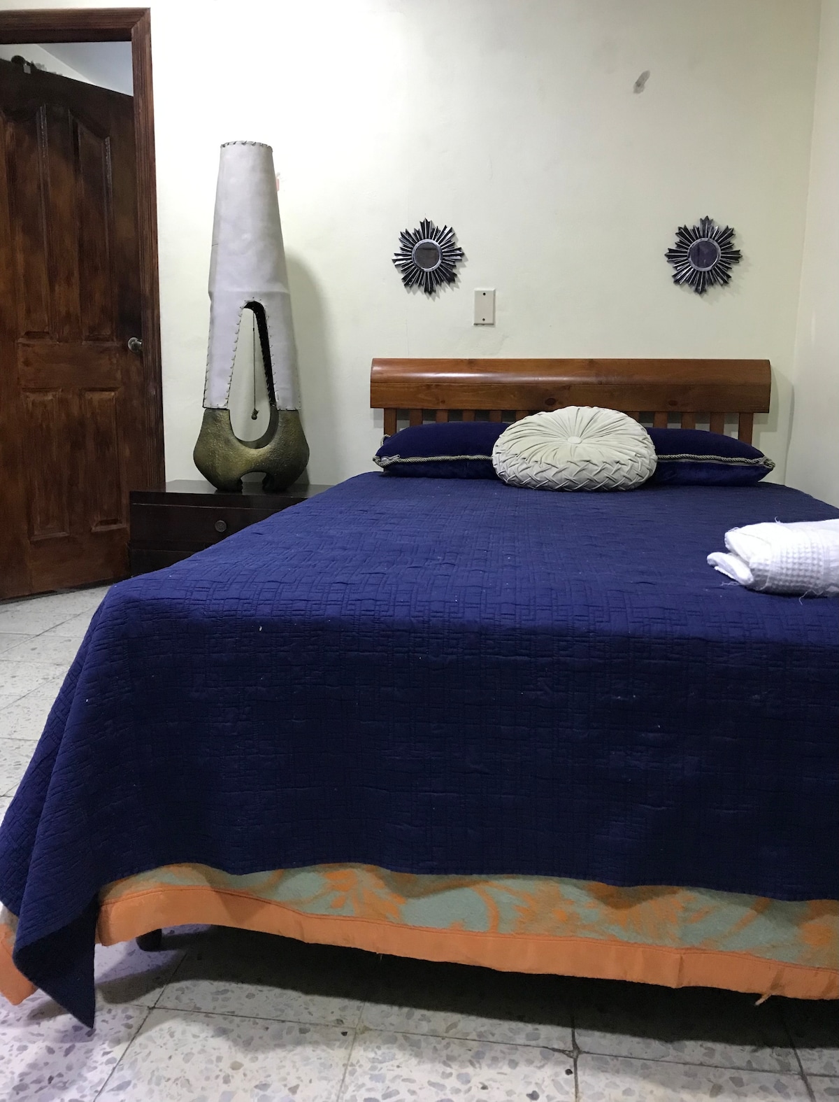 A spacious bed is dressed in a deep blue coverlet, complemented by decorative pillows. A bedside table stands nearby, along with a tall, unique lamp. Wall art features circular designs, while a wooden door provides access to the room.