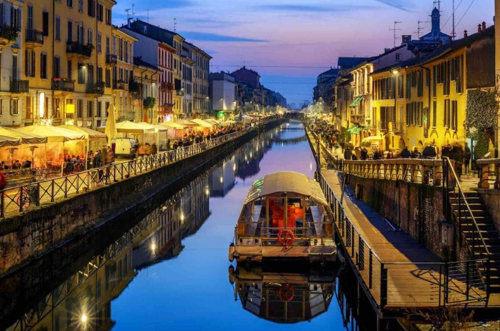 A tranquil evening scene along the Navigli, showcasing illuminated buildings lining the canal. Reflections of warm lights dance on the water's surface, while a boat is moored along the wooden walkway, adding to the serene ambiance of the location.