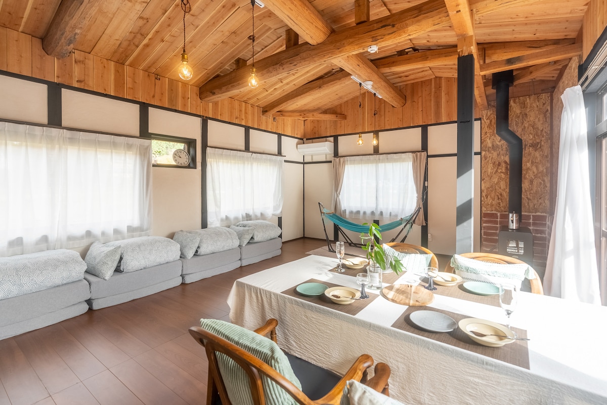 A spacious interior features wooden beams and soft lighting. A large dining table is set with dishes, while comfortable seating options, including a hammock, create a relaxed atmosphere. Sunlight filters through sheer curtains, illuminating the inviting space.