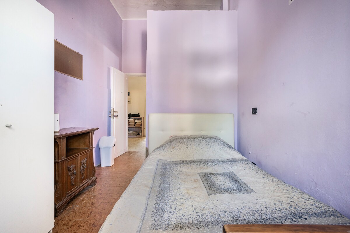 A spacious sleeping area features soft purple walls. A large bed is dressed with a light-patterned coverlet and placed against the wall. A wooden dresser offers storage, while a bathroom door is visible in the background.