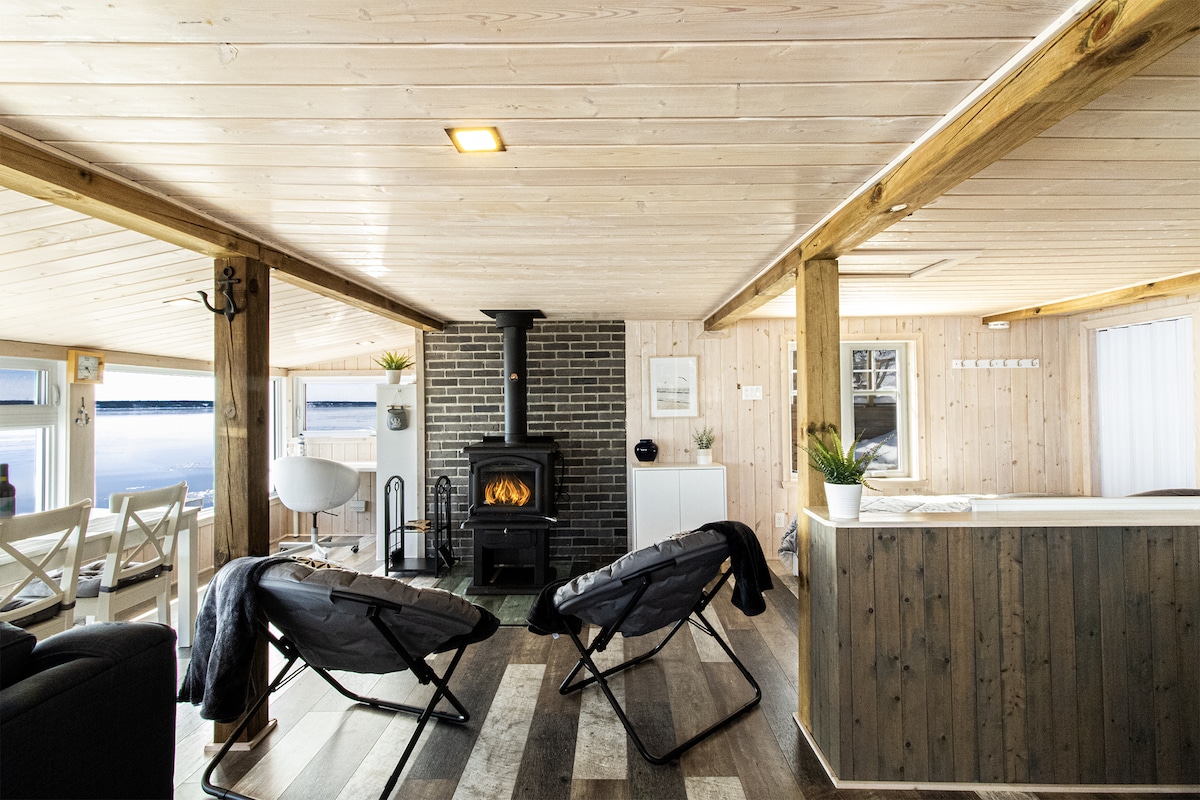 A spacious open-plan interior features wooden walls and a cozy wood stove. Two lounge chairs are positioned facing the water, while large windows ensure ample natural light and views of the river. The decor is minimal yet warm, highlighting a relaxing atmosphere.