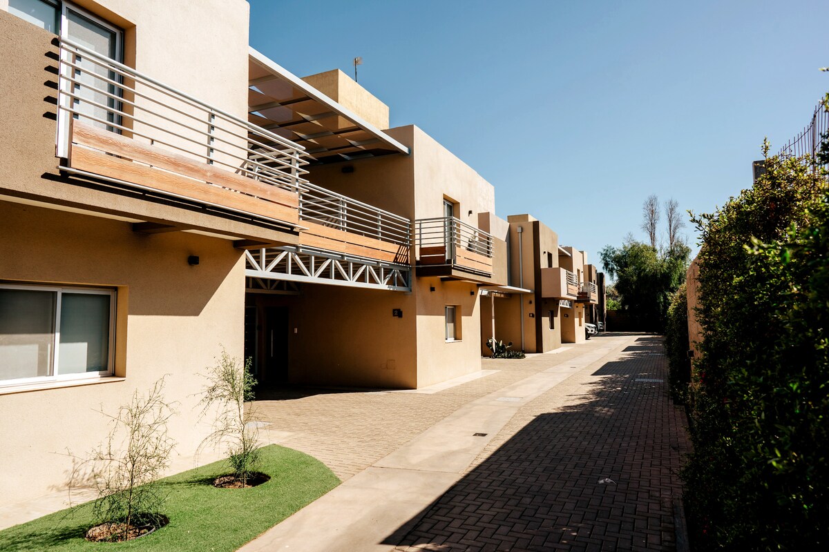 The exterior of the 'Portal del Aguaribay' complex is shown, featuring a broad pathway lined with twelve modern units. Each unit has a balcony and large windows, allowing natural light to flow in. The surrounding landscaping includes small shrubs, enhancing the inviting atmosphere.