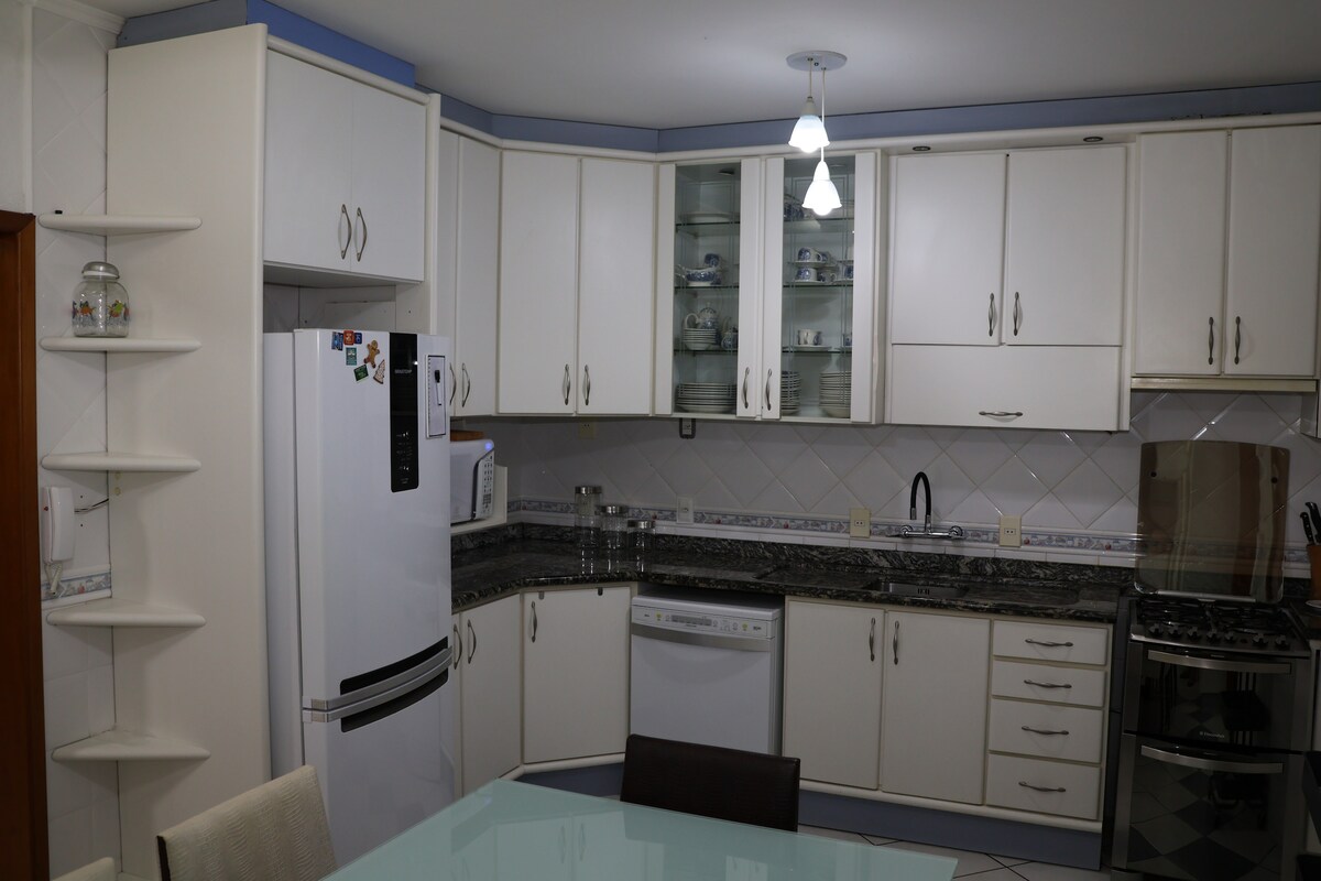The kitchen features white cabinetry with glass-front shelves. A refrigerator, microwave, and dishwasher are included. A stainless steel gas stove is positioned adjacent to a granite countertop. A dining table with chairs occupies the center of the space, with overhead lighting providing illumination.