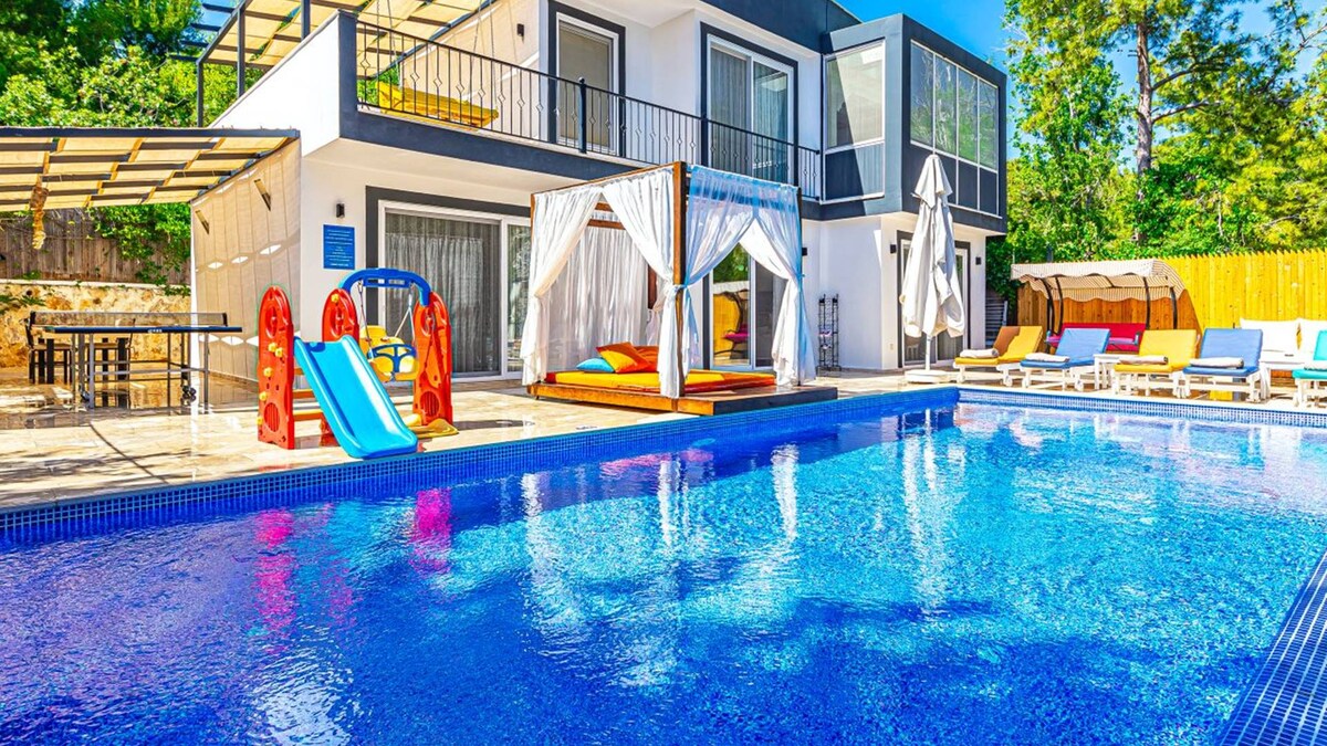 A spacious outdoor area features a large swimming pool with a vibrant blue surface. Sun loungers are arranged around the pool, with a shaded seating area and colorful play equipment visible for children.