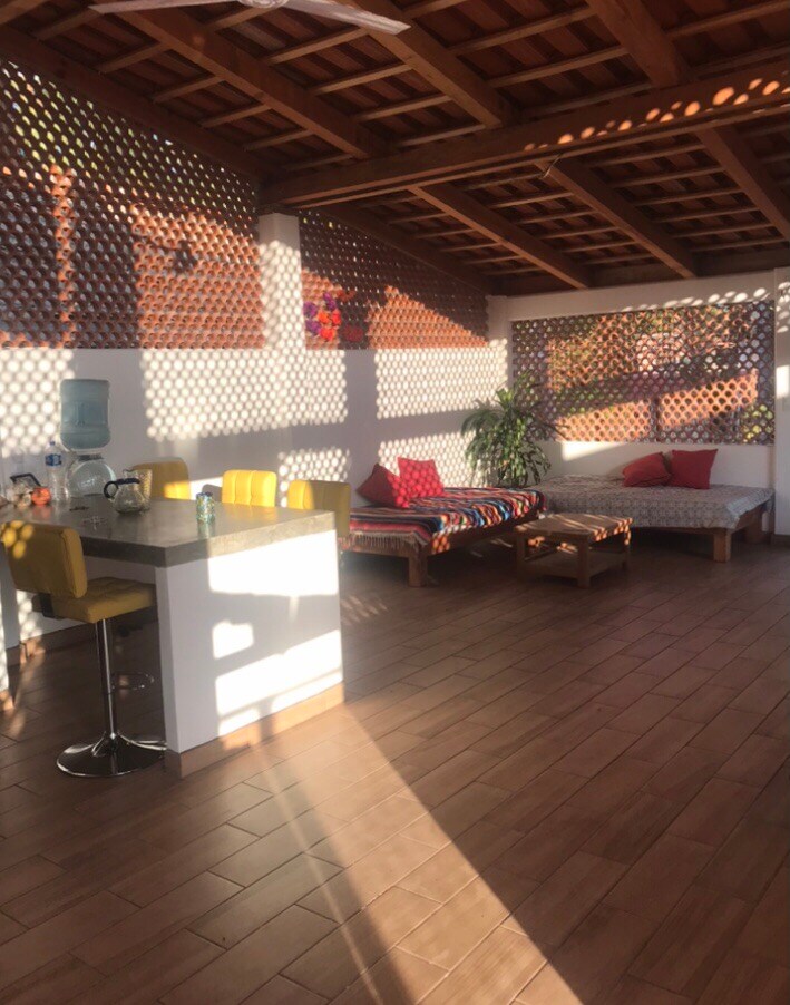 A well-lit common area features wooden ceiling beams and a tile floor. Comfortable seating with cushions is provided along the walls, while a bar area with yellow stools is visible. Natural light creates patterns on the surfaces from the lattice design of the walls.