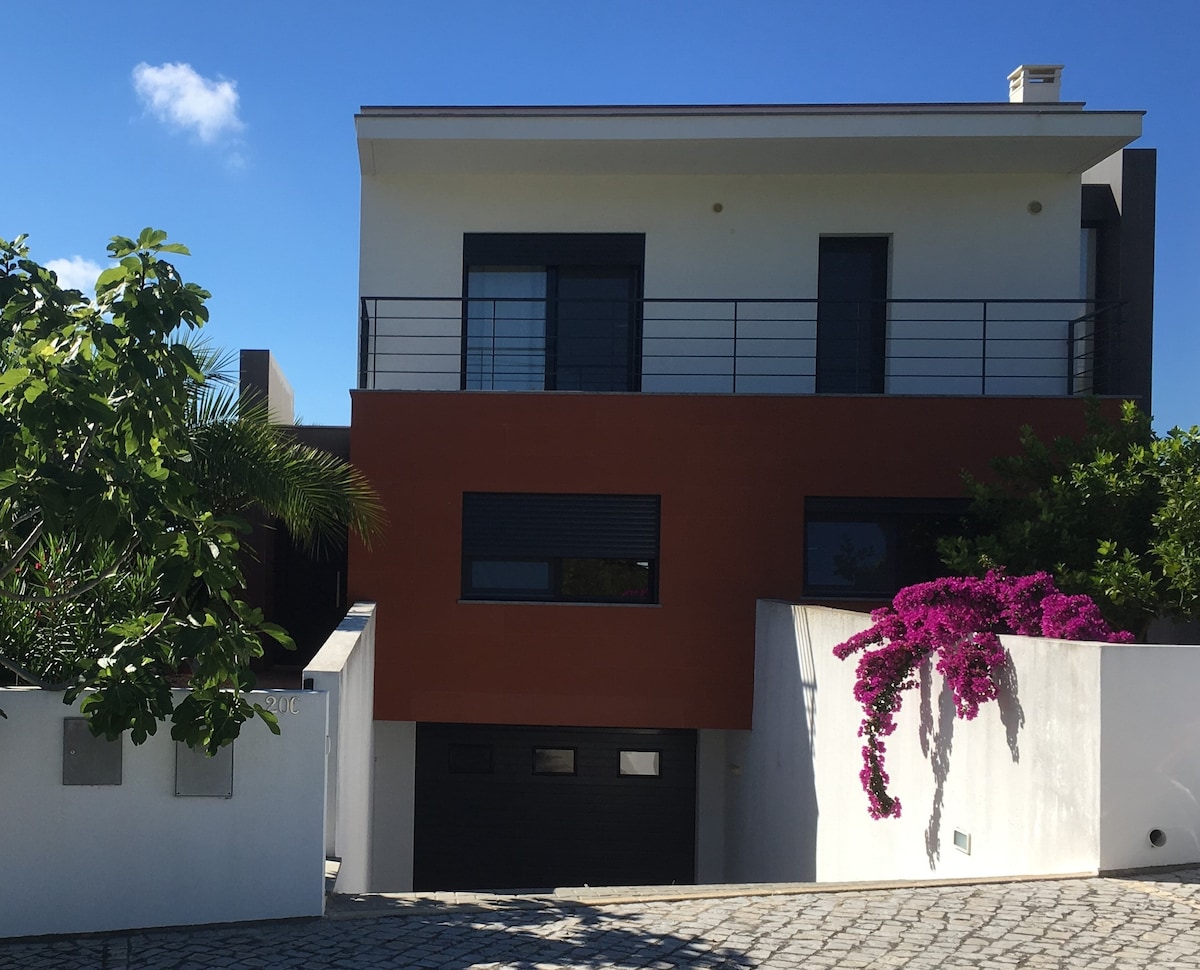 The modern two-story house features a blend of vibrant orange and white exterior with large windows. Lush greenery and blooming bougainvillea add color to the facade. A paved pathway leads to the main entrance, enhancing the welcoming appearance.