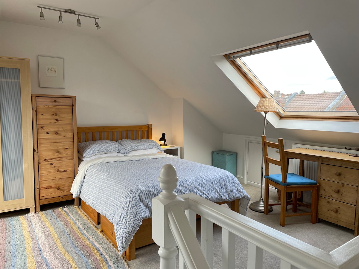A loft room features a generous layout with a spacious bed and a desk accompanied by a chair. Natural light floods in through a Velux window, illuminating the wooden furniture, including a wardrobe and storage chest. A colorful rug adds warmth to the neutral carpeted floor.