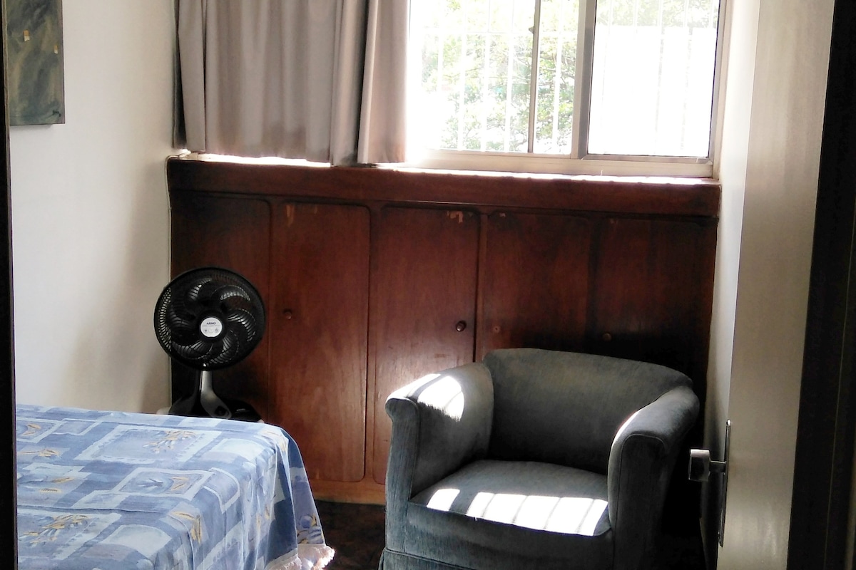 A single room is presented with a spacious bed covered in a blue patterned blanket. A comfortable armchair is positioned near a large window, which allows natural light to fill the room. A standing fan is visible beside the bed.