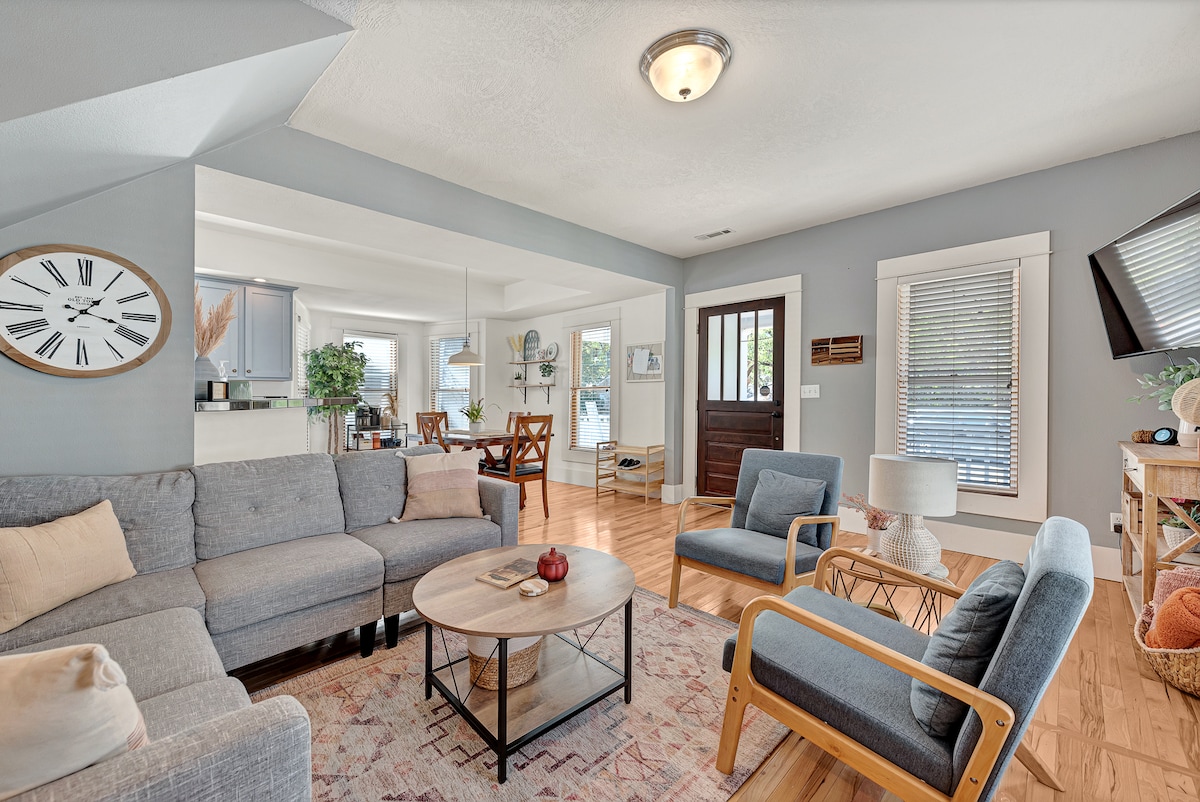 A welcoming living area features a comfortable gray sectional sofa and a round coffee table at its center. Light wood flooring contrasts with the soft hues of the decor. A large wall clock and a cozy accent chair add warmth, while natural light streams through the window.