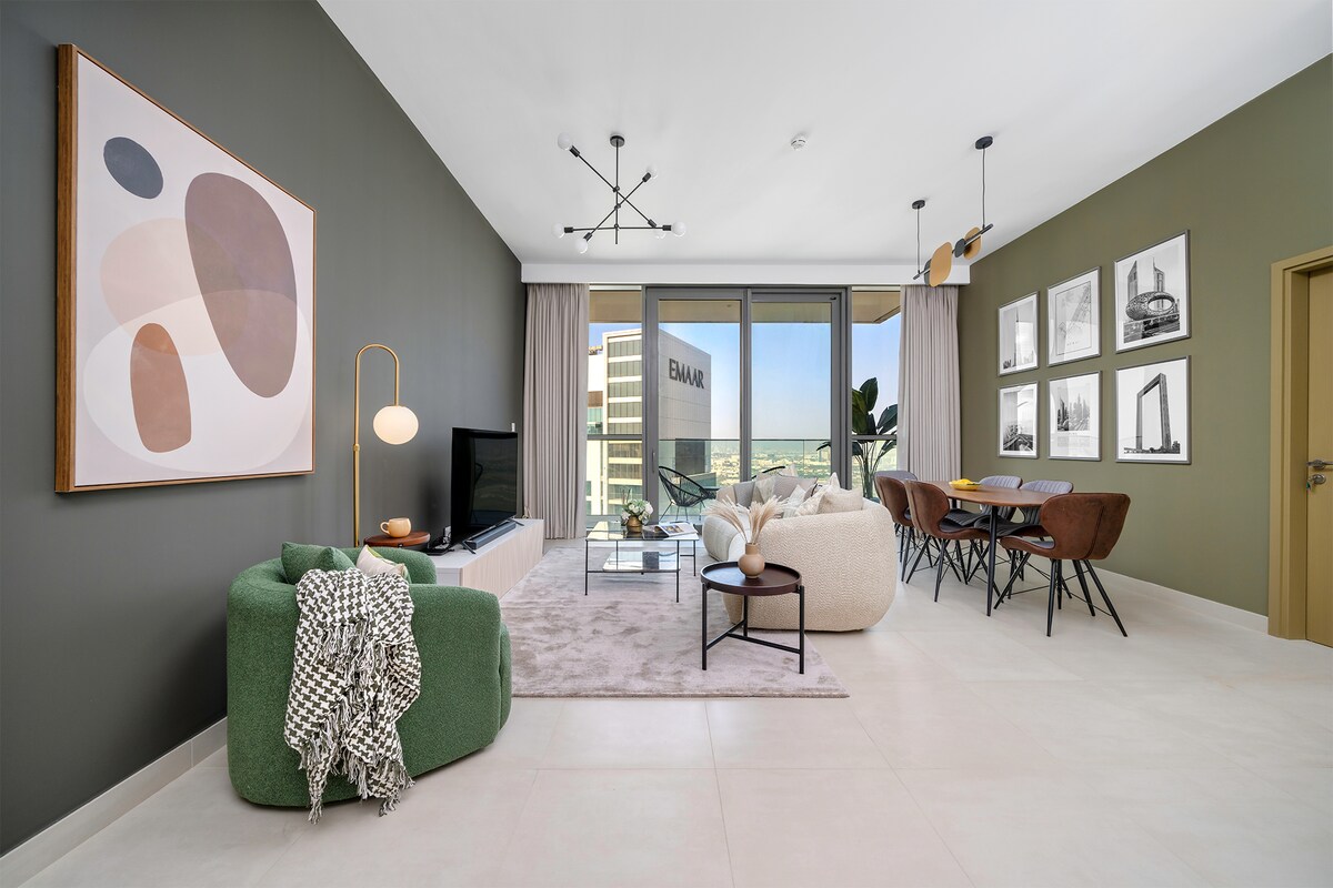 A modern living area is highlighted by an abstract art piece on one wall, with a flat-screen television positioned nearby. Natural light floods through large glass doors, leading to a balcony. A stylish dining table and cozy seating arrangements enhance the inviting atmosphere.