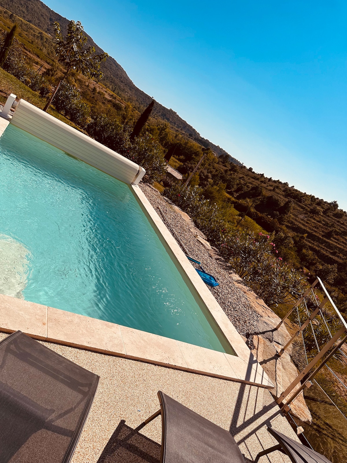 A private swimming pool features clear blue water, surrounded by landscaped greenery and rolling hills. Two lounge chairs are positioned nearby on a patio surface, providing an inviting space for relaxation under the sunny sky.